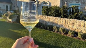 Un verre transparent sur lequel sont inscrits les mots « De Coupage » et « Chardonnay », placé devant une maison en pierre dotée d’un mur de pierre et agrémentée de verdure.