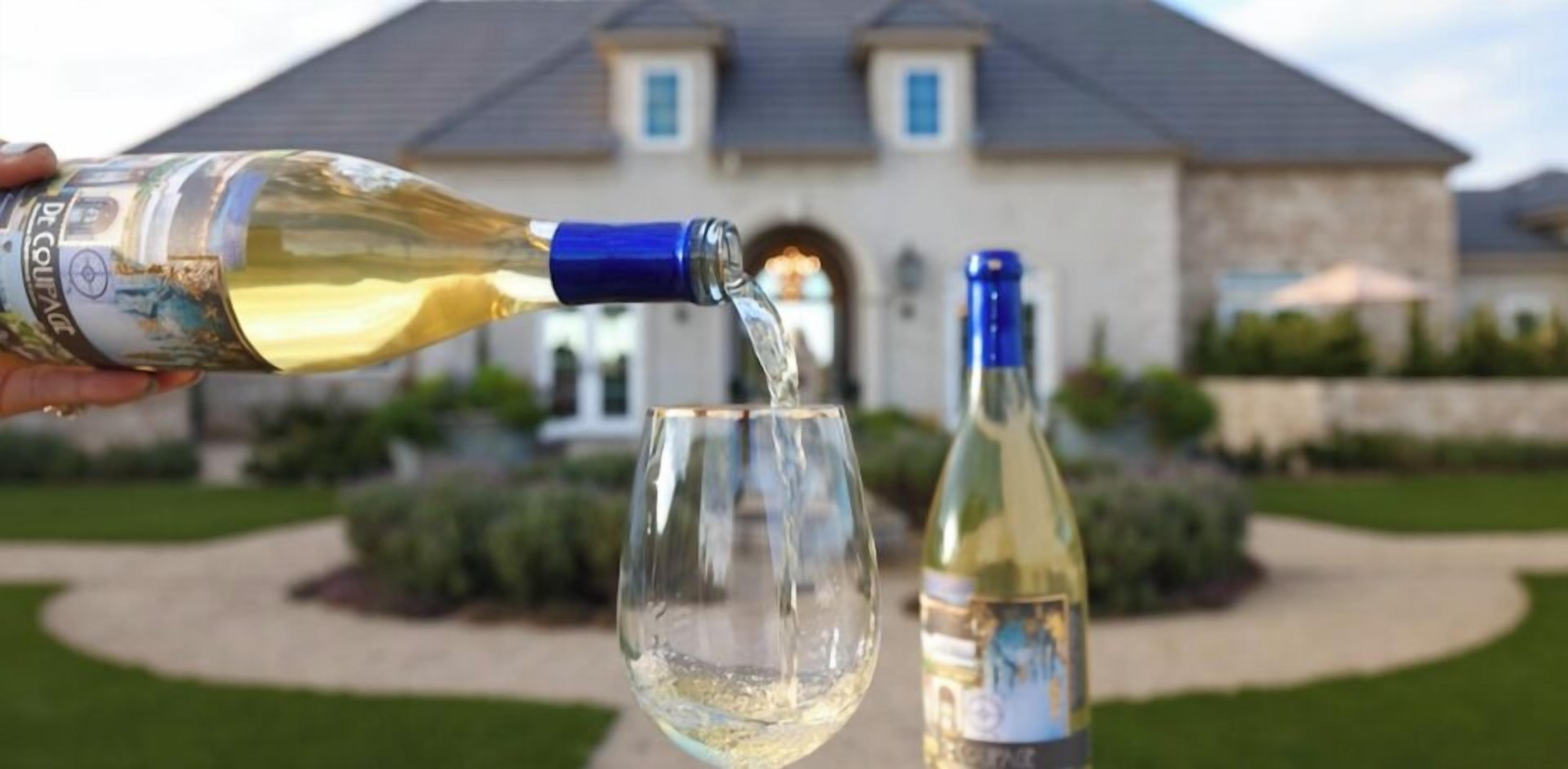 Une bouteille de vin est versée dans un verre, avec une maison en arrière-plan.