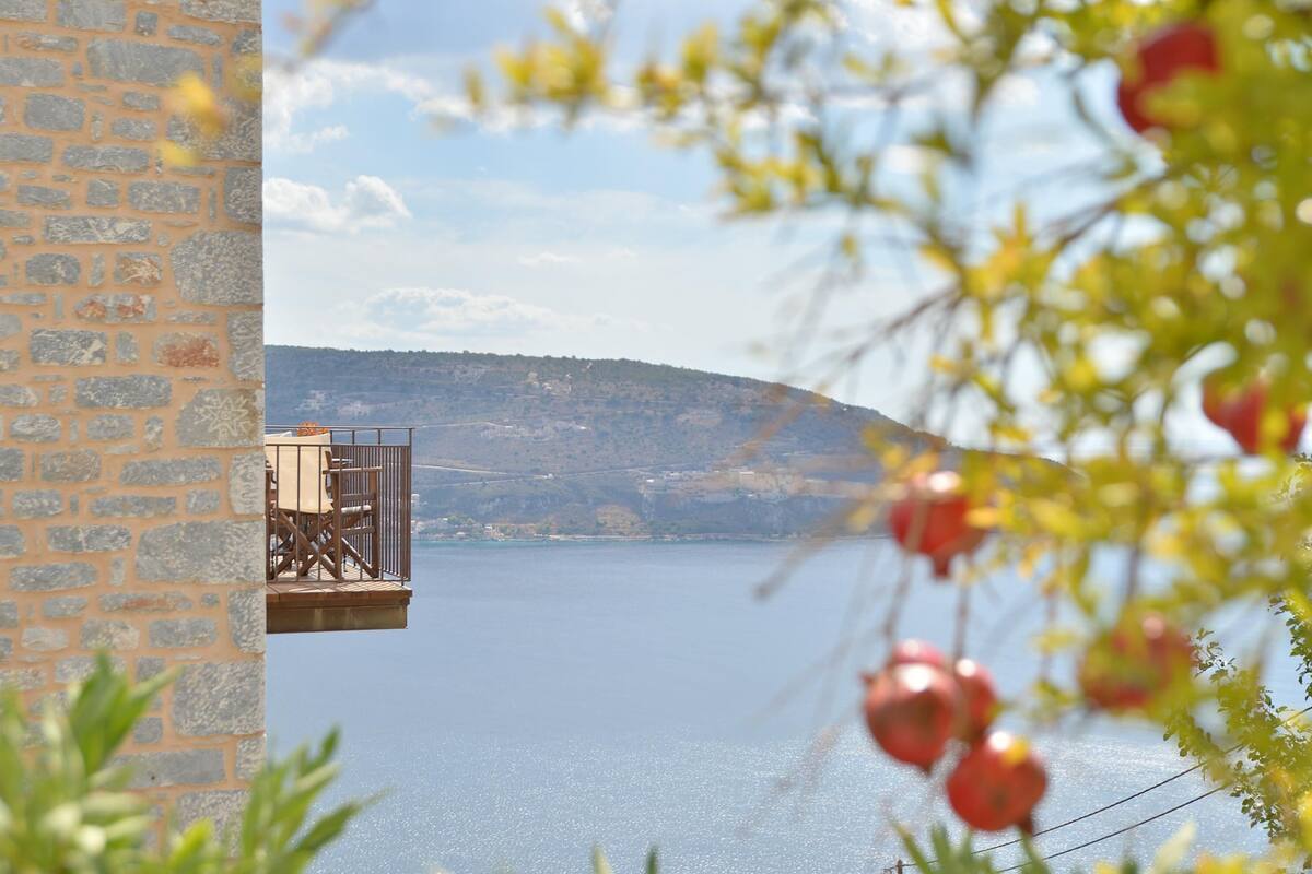 A stone building with a balcony overlooking a calm body of water and a hilly landscape.
