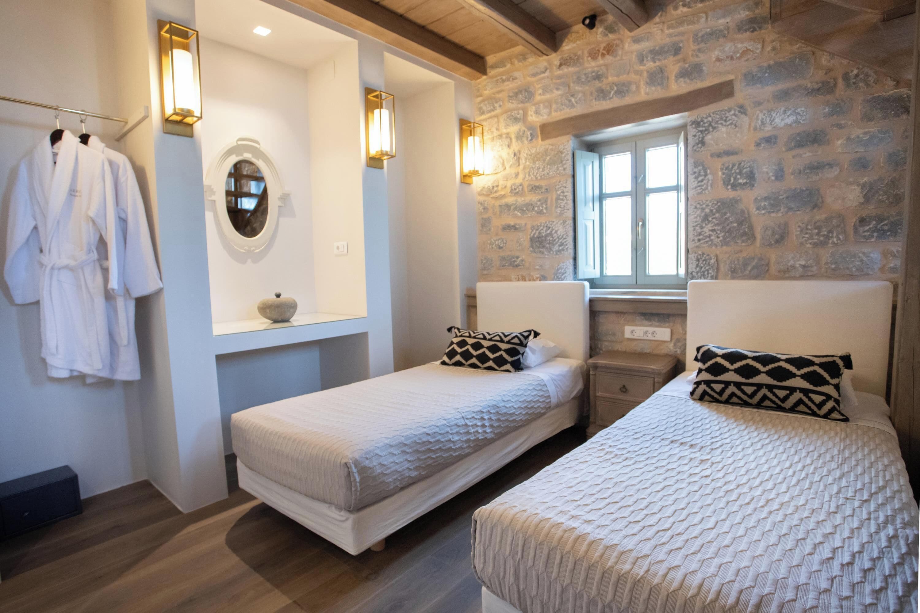 Une chambre d’hôtel avec deux lits simples, un porte-peignoir, un miroir et des éléments décoratifs en pierre sur les murs.