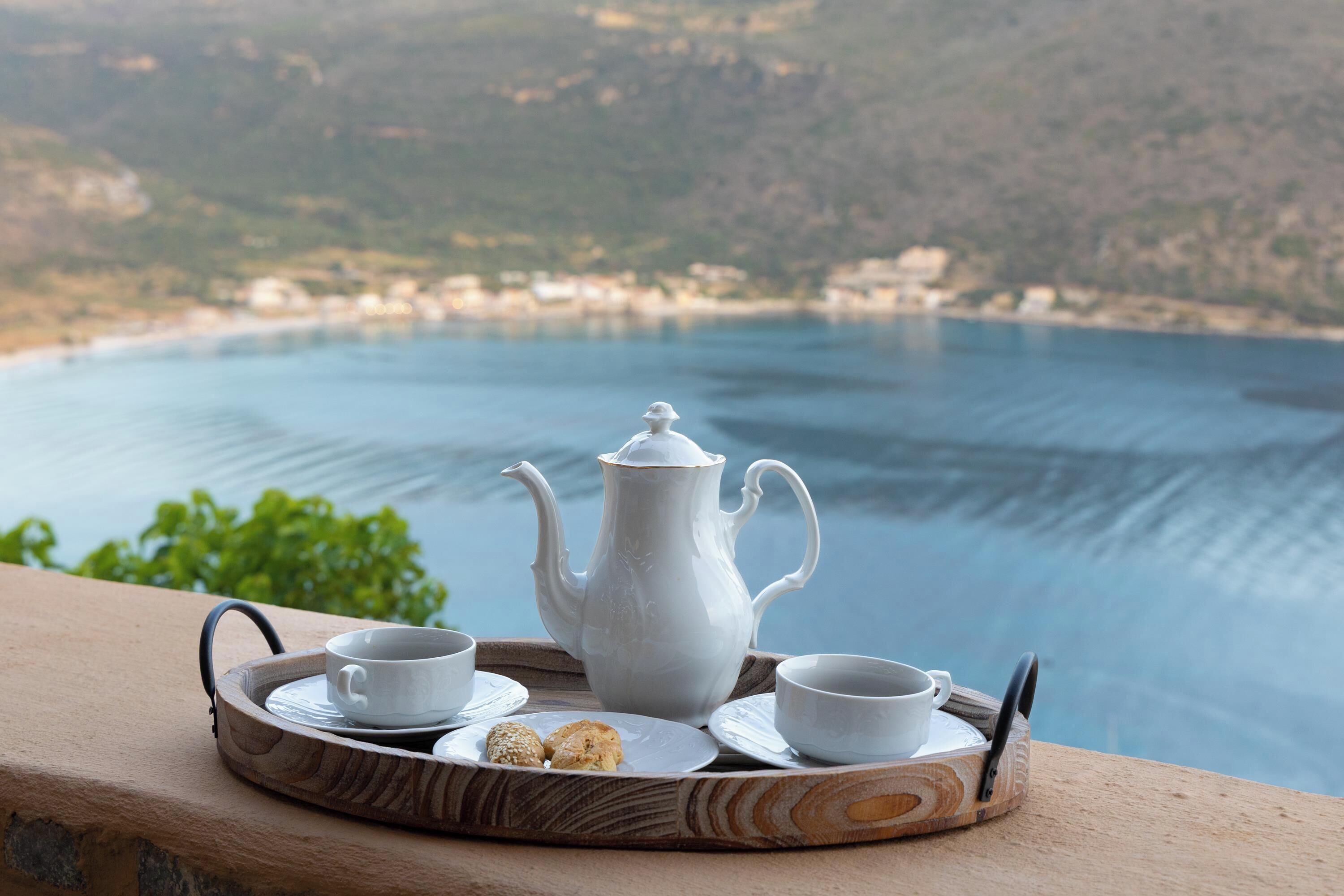 A tray with a teapot, cups, and a biscuit on a balcony overlooking a body of water.