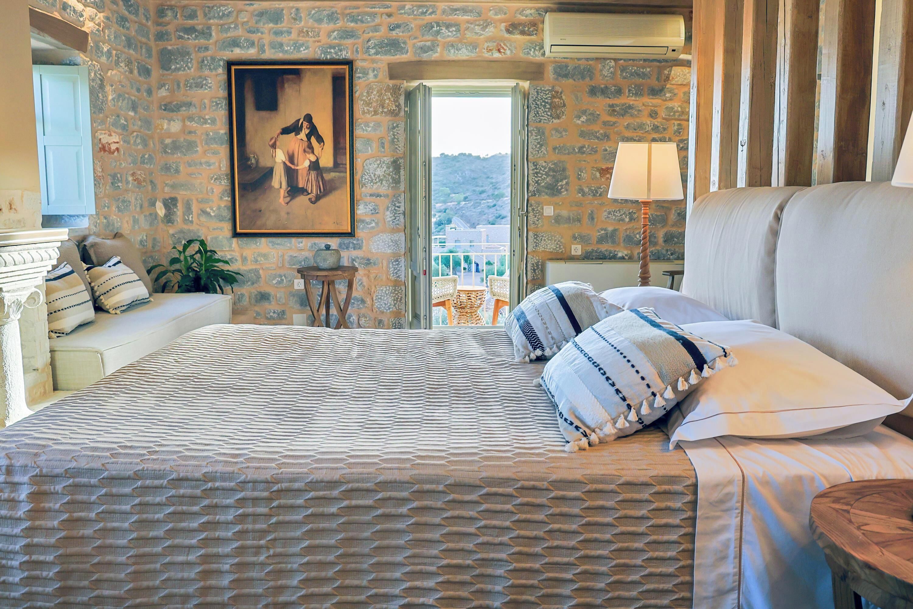A bedroom with a stone wall, a painting, a bed with patterned bedding, a nightstand, a lamp, and a view of the outdoors.