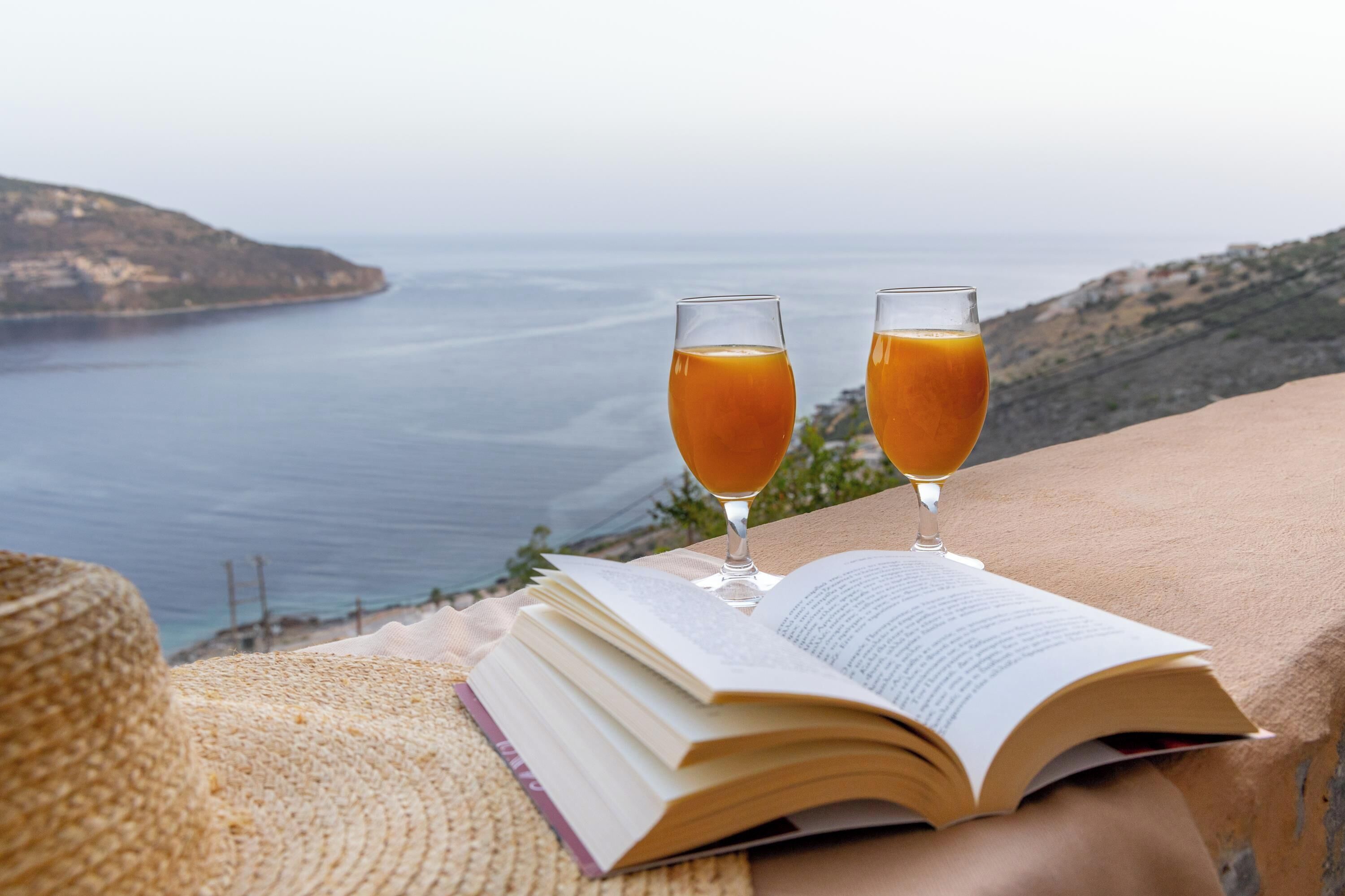 Un llibre obert sobre una taula amb dos vasos de suc de taronja, amb vista a un paisatge costaner.
