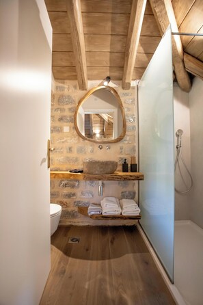 A bathroom with a stone wall, wooden beams, a wooden shelf with a bowl sink, a mirror, a toilet, and a glass shower enclosure. - Akrolithi Guesthouse (East Mani)