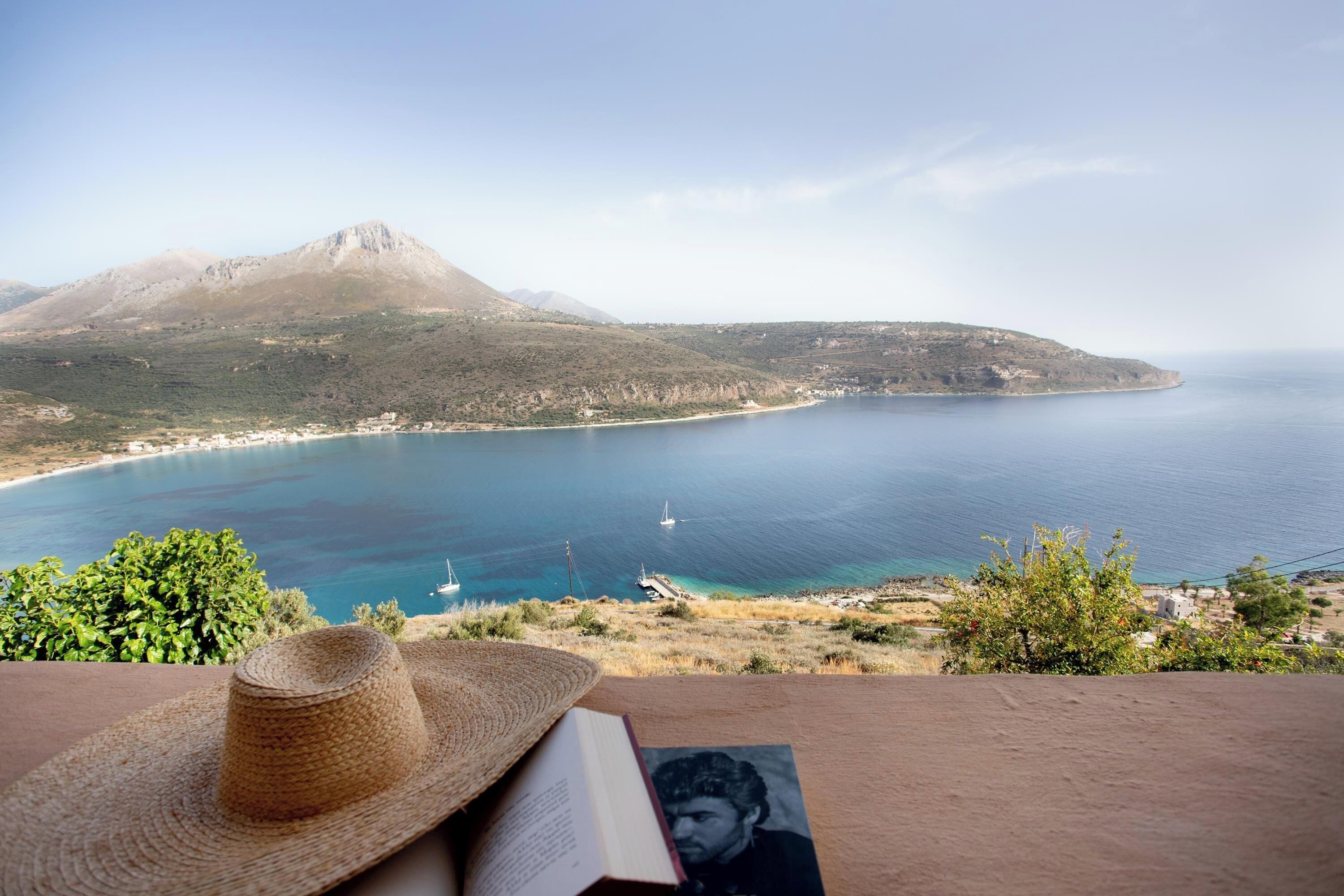 Une vue d’un paysage côtier avec une montagne, une plage et un livre, accompagné d’un chapeau de paille posé sur une table.