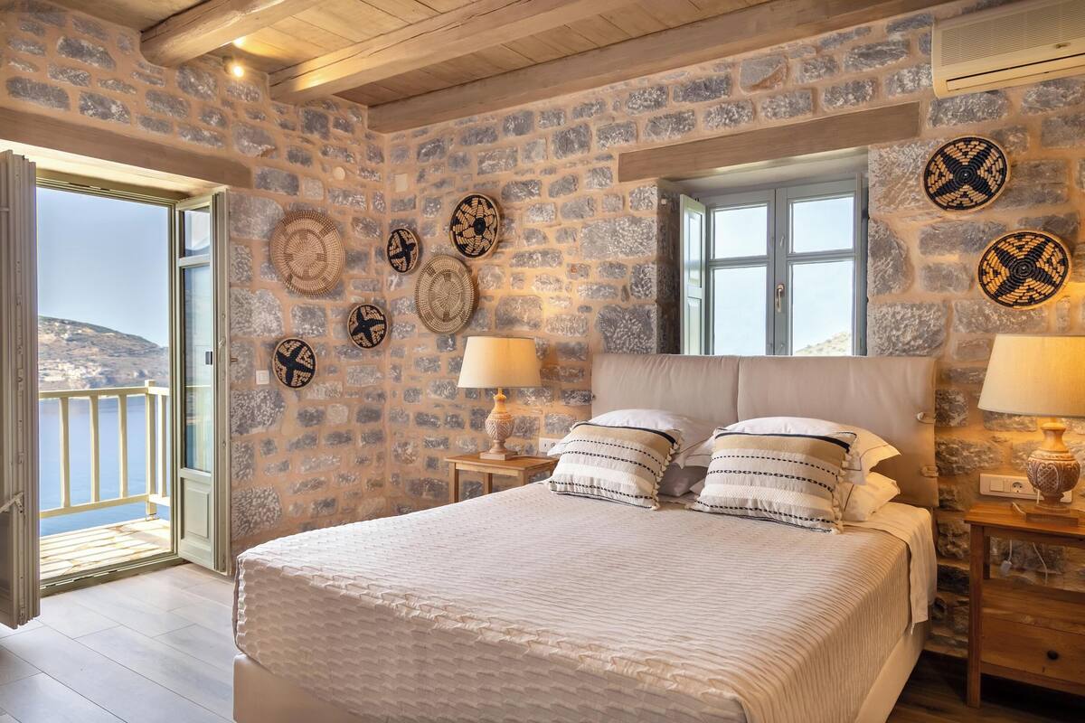 A stone-walled bedroom with a bed, two bedside tables with lamps, and a view of the sea through an open door.