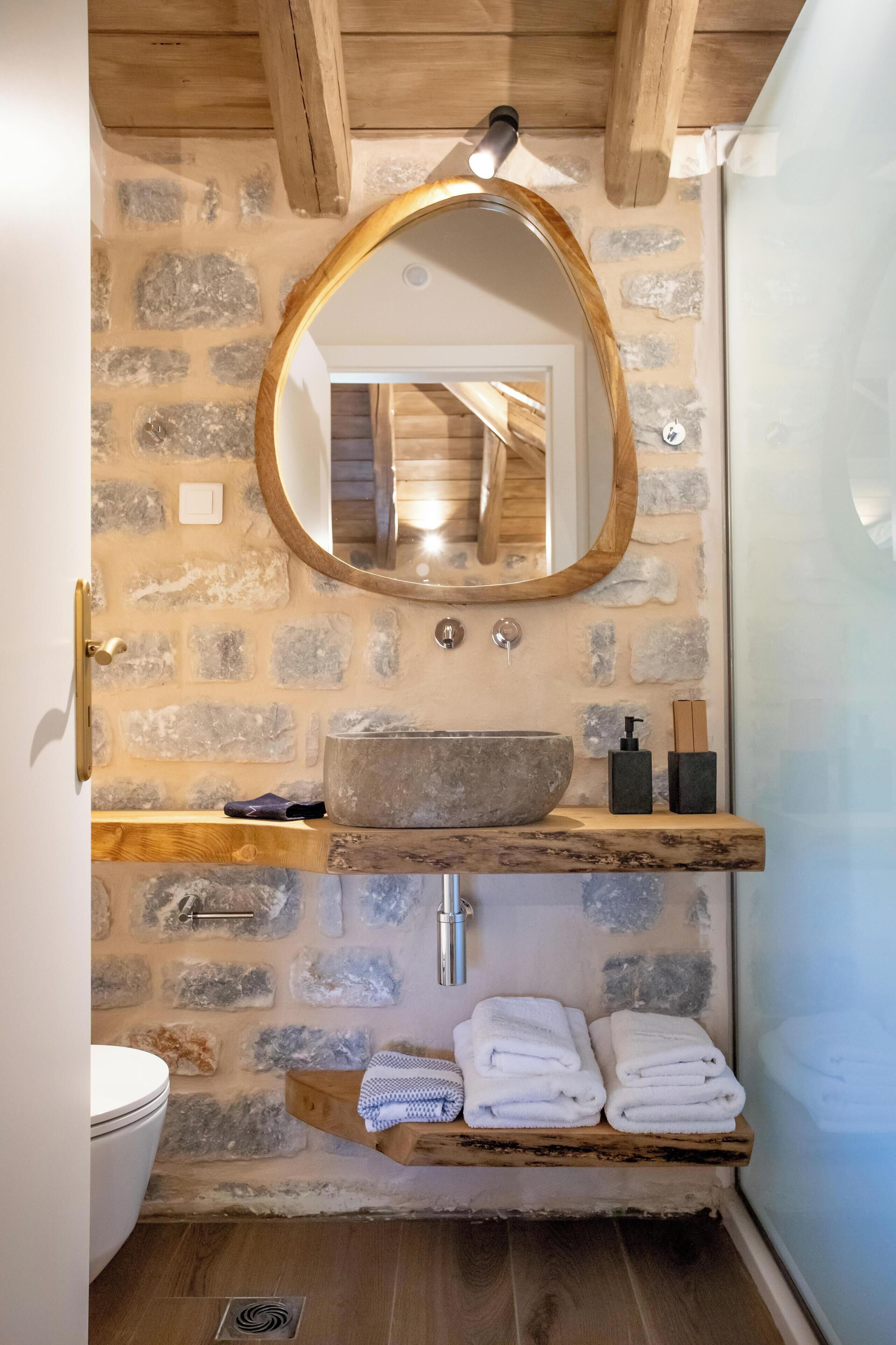 A bathroom with a stone wall, wooden beams, a large oval mirror, a stone sink, and neatly folded towels on a shelf.