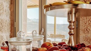 Une table de buffet avec des pommes et des bananes, une chaise en osier et une vue sur les montagnes à travers de grandes fenêtres.