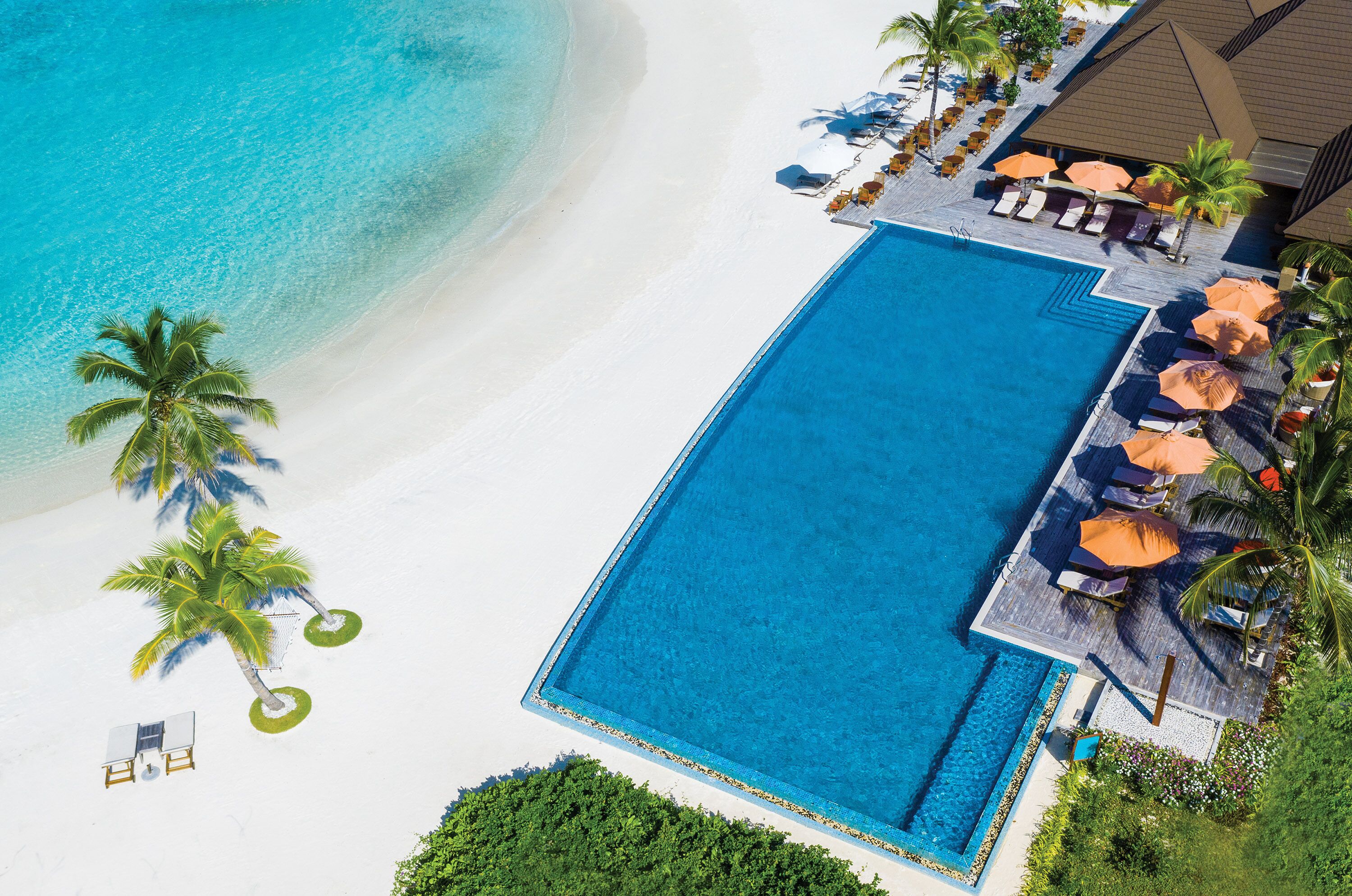Piscina de un resort con agua azul cristalina, rodeada de sillas de descanso y sombrillas, junto a una playa de arena con palmeras.