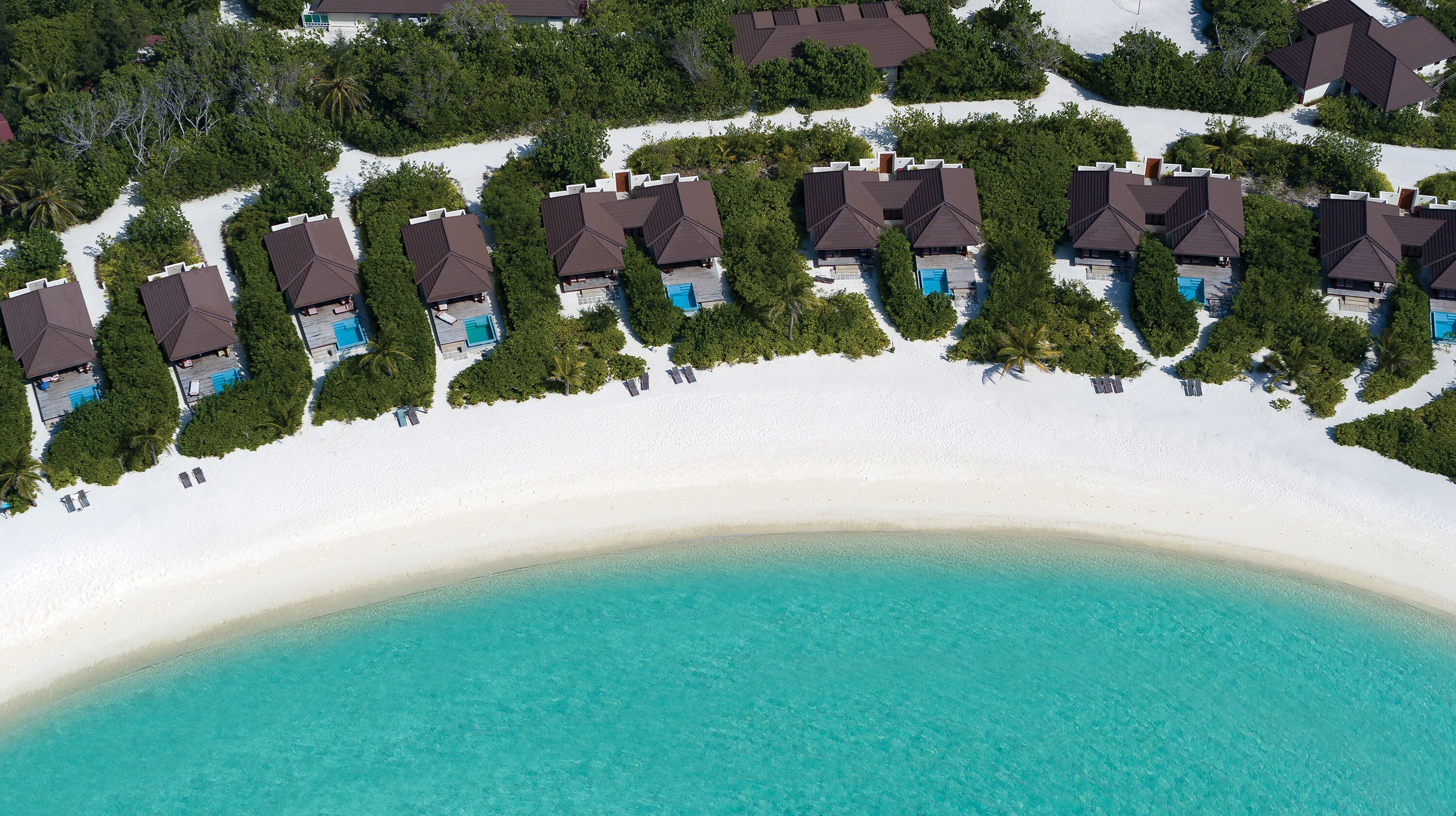 An aerial view of a resort with multiple bungalow-style buildings, each with a private pool, situated on a white sandy beach with clear turquoise waters.