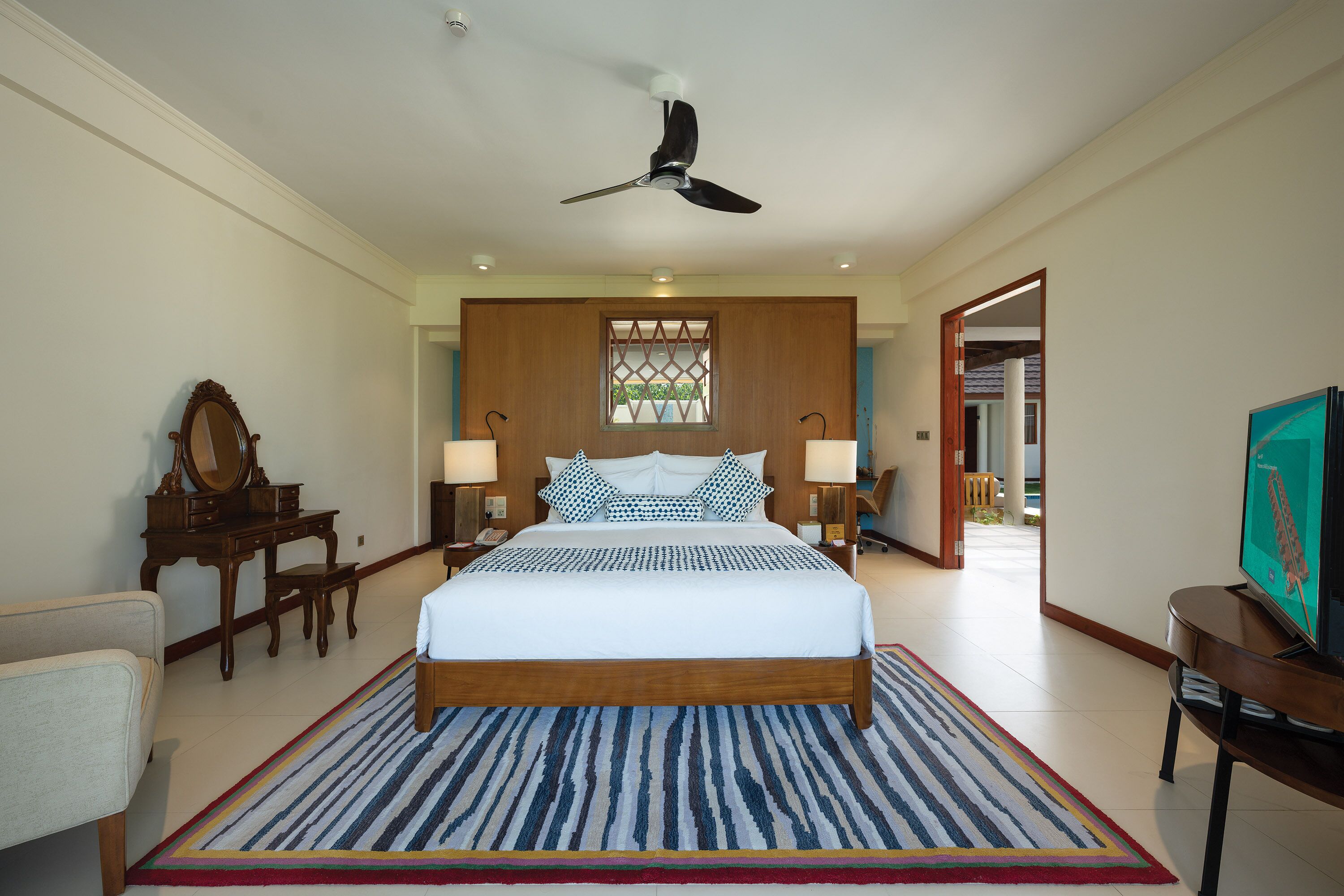 A bedroom with a large bed, wooden headboard, bedside lamps, a desk with a mirror, and a television on a wooden stand.