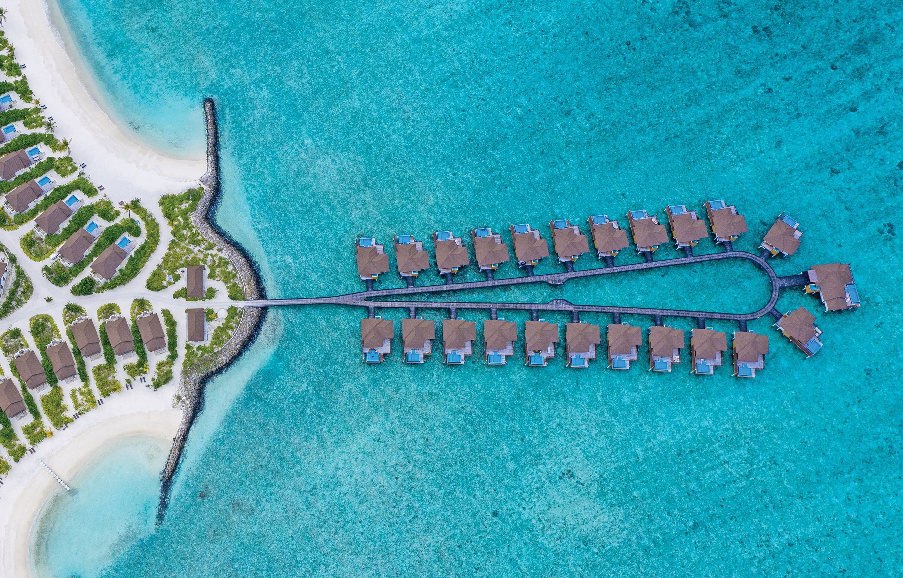 An aerial view of a resort with overwater bungalows connected by a pier, a beach area with lounge chairs, and a clear turquoise sea.