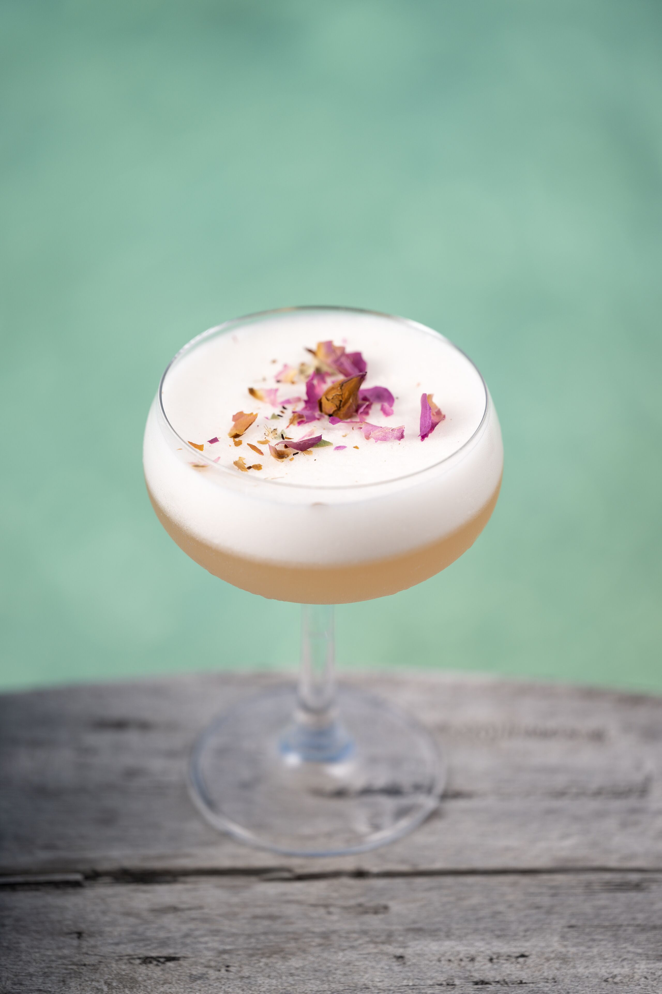 A cocktail in a stemmed glass with a creamy top, garnished with petals and spices, on a wooden surface against a green backdrop.