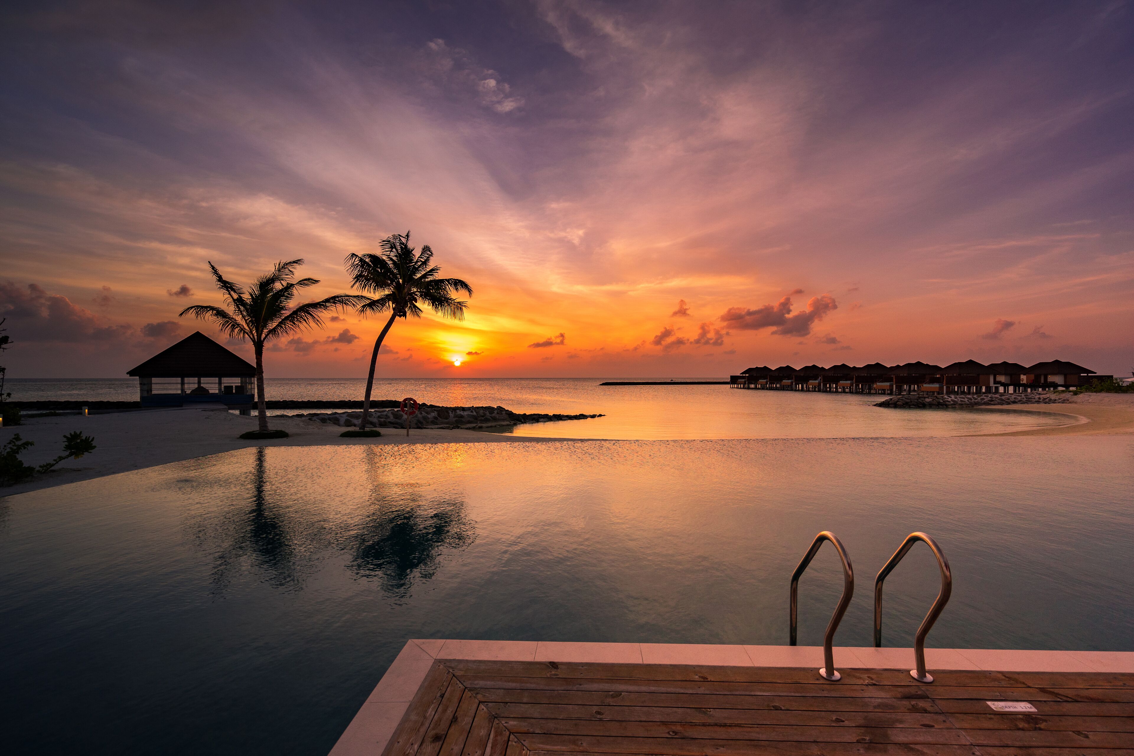 A serene sunset over a calm sea with a wooden deck and two palm trees.
