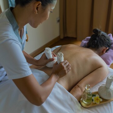 A massage therapy session in a room with a bed, a massage table, and a window with curtains.