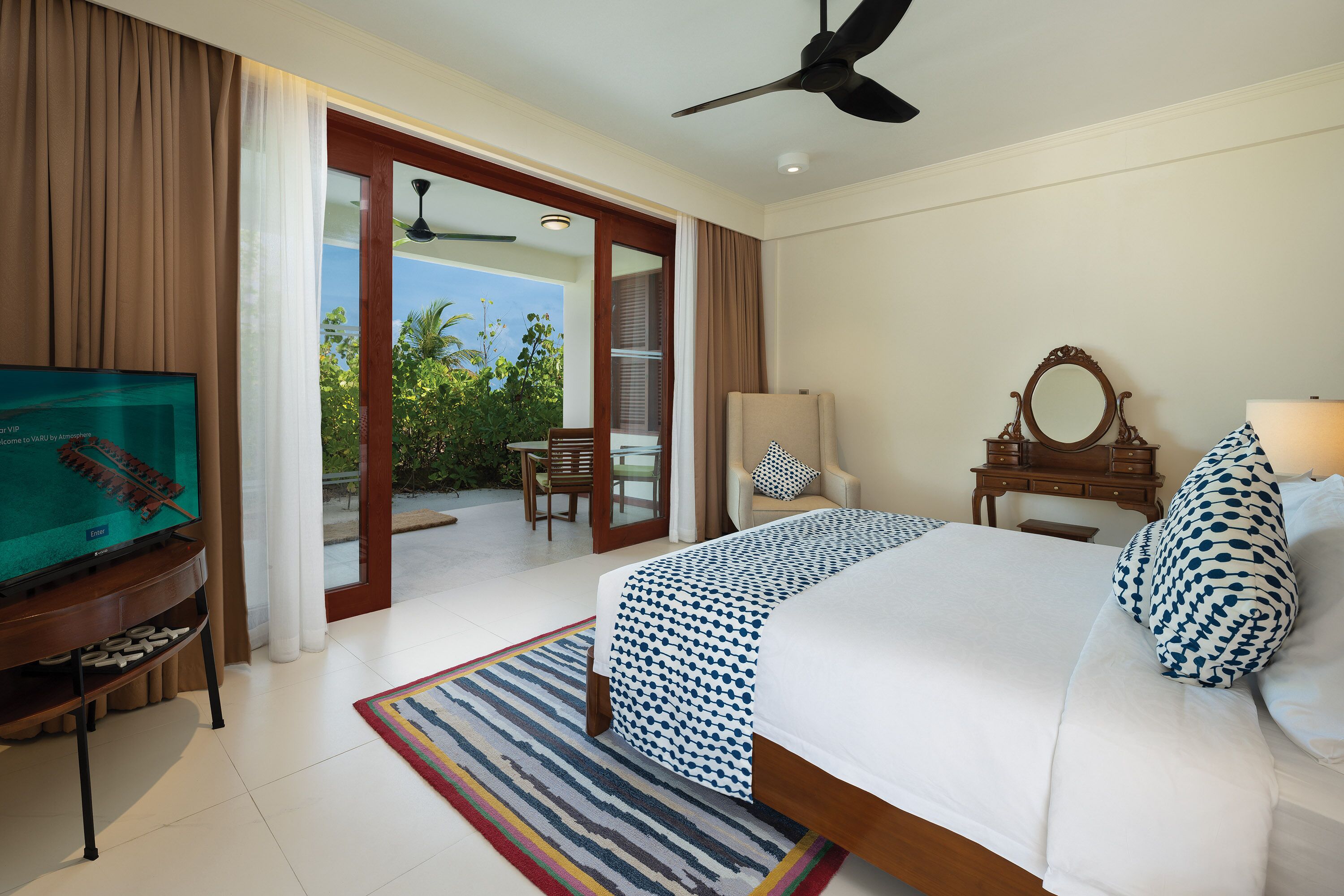 A hotel room with a bed, a television, a ceiling fan, a window with curtains, a chair, and a view of greenery outside.