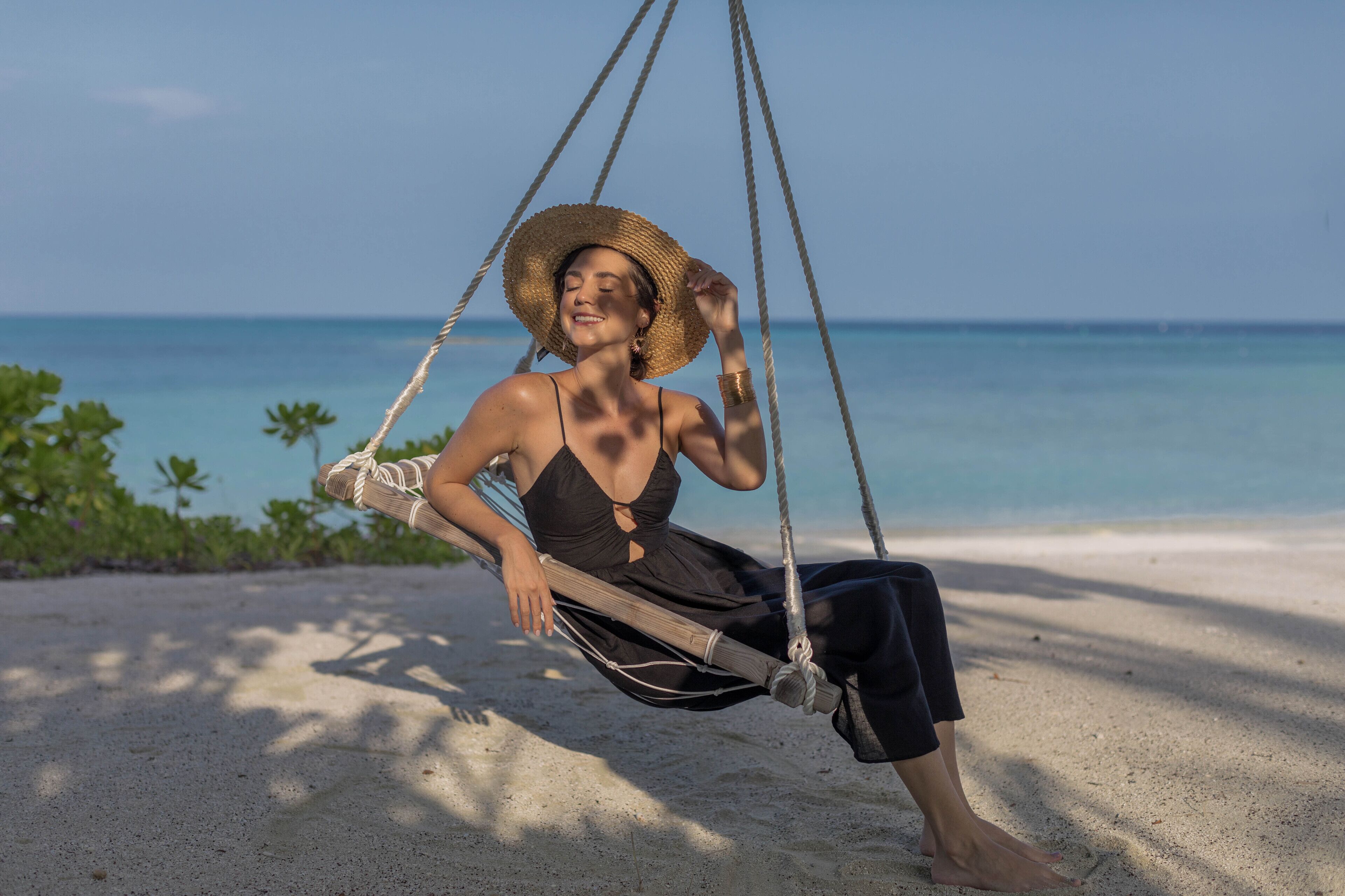 A person sitting on a hammock swing on a sandy beach with a clear view of the ocean.