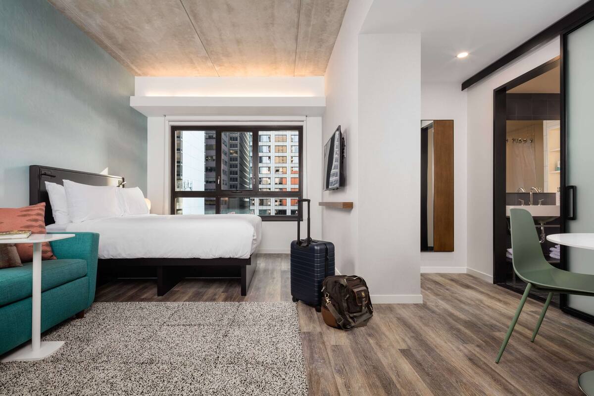 A modern hotel room with a bed, a small table, a sofa, a suitcase, and a bathroom visible through an open door.