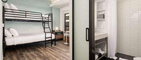 A room with a bunk bed, a desk, a chair, a small table, and a bathroom with a shower and toilet.