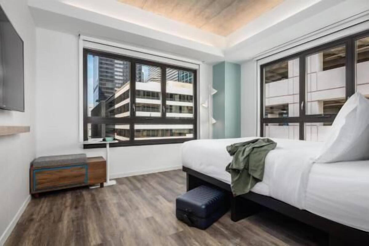 A modern bedroom with a bed, a window with a view of buildings, a small table, and a bench.