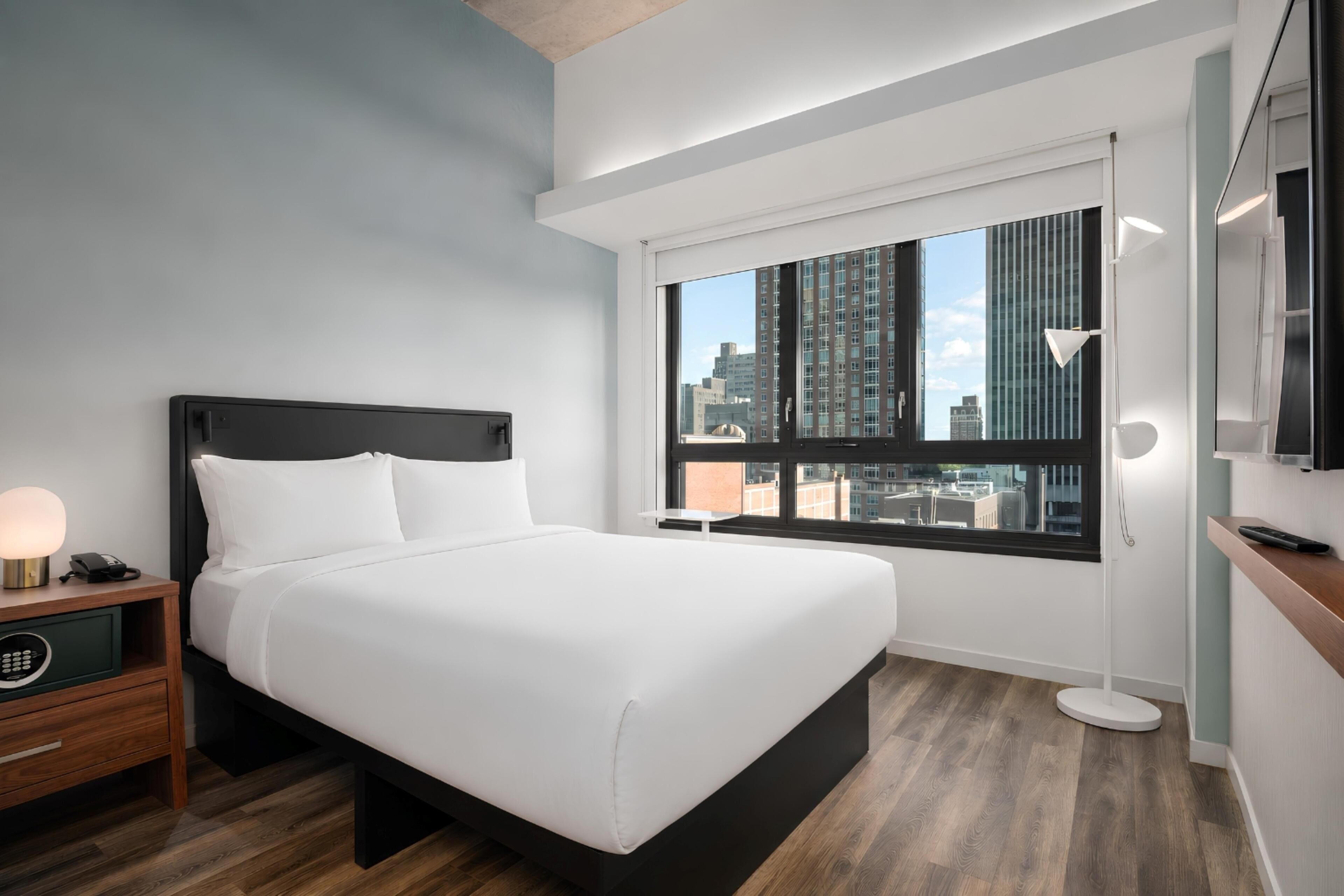 A hotel room with a large bed, a nightstand with a lamp, a TV, and a window with a city view.
