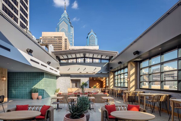 A rooftop bar with outdoor seating, large windows, and a view of the city skyline.