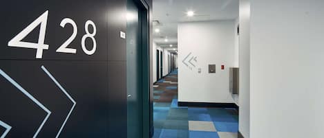 Un pasillo moderno de hotel con alfombra a cuadros, un letrero con número en la pared y un pasillo que conduce a varias habitaciones.