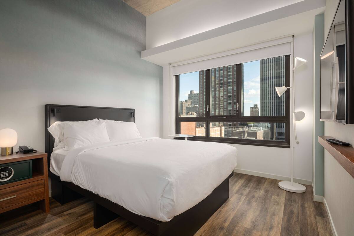 A modern hotel room with a large bed, a nightstand with a lamp, a bedside table, and a view of the city through a large window.