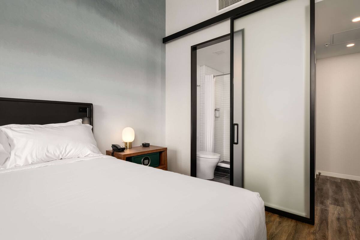 A hotel room with a bed, a nightstand with a lamp, a wooden side table, a sliding door to the bathroom, and a view of the bathroom through the door.