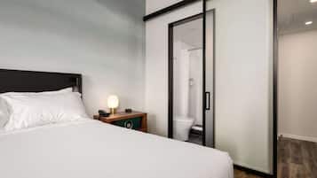 A hotel room with a bed, a nightstand with a lamp, a wooden side table, a sliding door to the bathroom, and a view of the bathroom through the door.