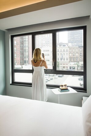 A woman in a white dress standing by a window with a view of city buildings.