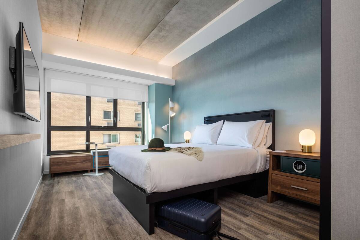 A modern hotel room with a large bed, bedside tables, a suitcase, and a view of the city through large windows.