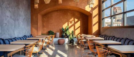 A modern cafe with wooden tables and orange chairs, large windows, and hanging wicker lamps.
