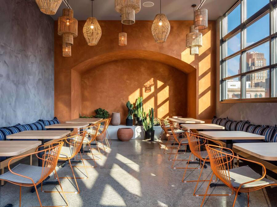 A modern cafe with wooden tables and orange chairs, large windows, and hanging wicker lamps.