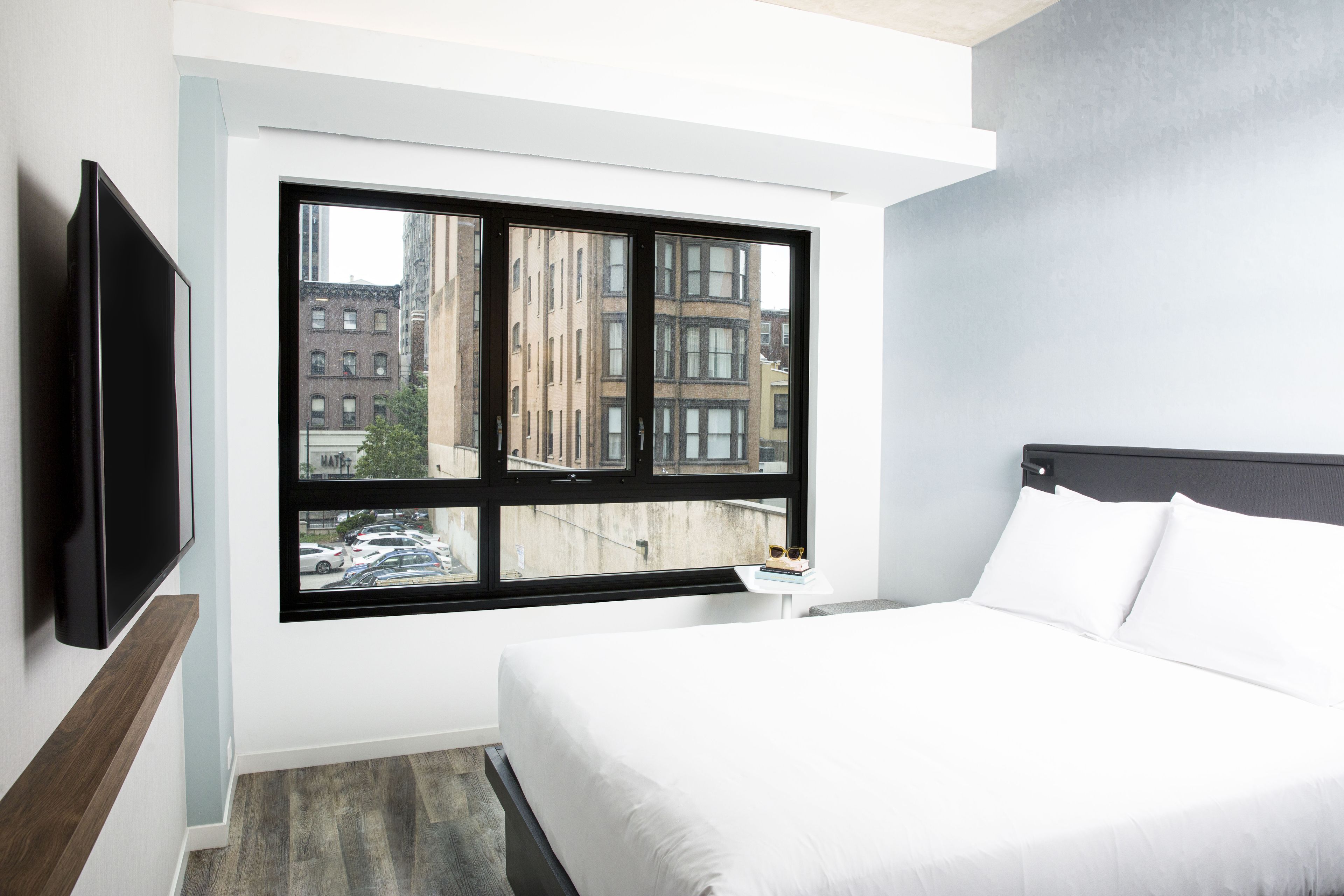 A modern hotel room with a large window, a flat-screen TV mounted on the wall, a neatly made bed with white linens, and a small bedside table with a book and glasses.