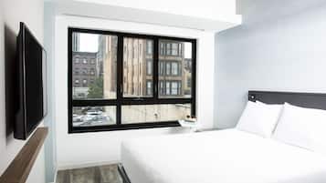 A modern hotel room with a large window, a flat-screen TV mounted on the wall, a neatly made bed with white linens, and a small bedside table with a book and glasses.