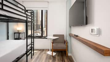 A hotel room with bunk beds, a small table, a chair, a TV, and a window with a city view.