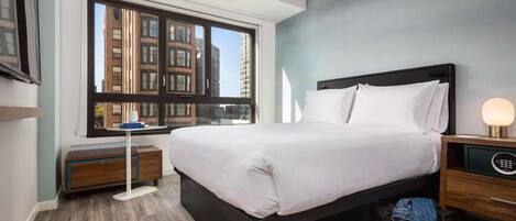 A modern hotel room with a large window, a bed, a bedside table with a lamp, a wooden nightstand, and a small wooden dresser.