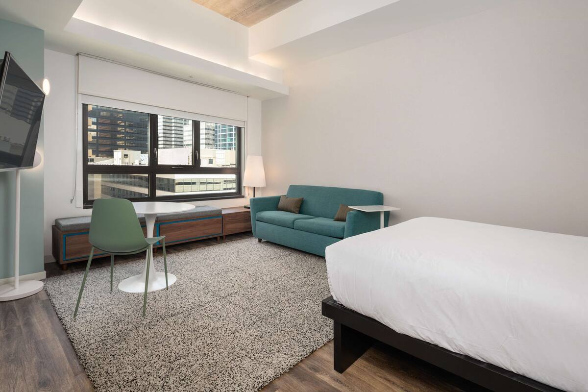 A modern hotel room with a bed, a green sofa, a small table, a chair, a desk, and a TV.