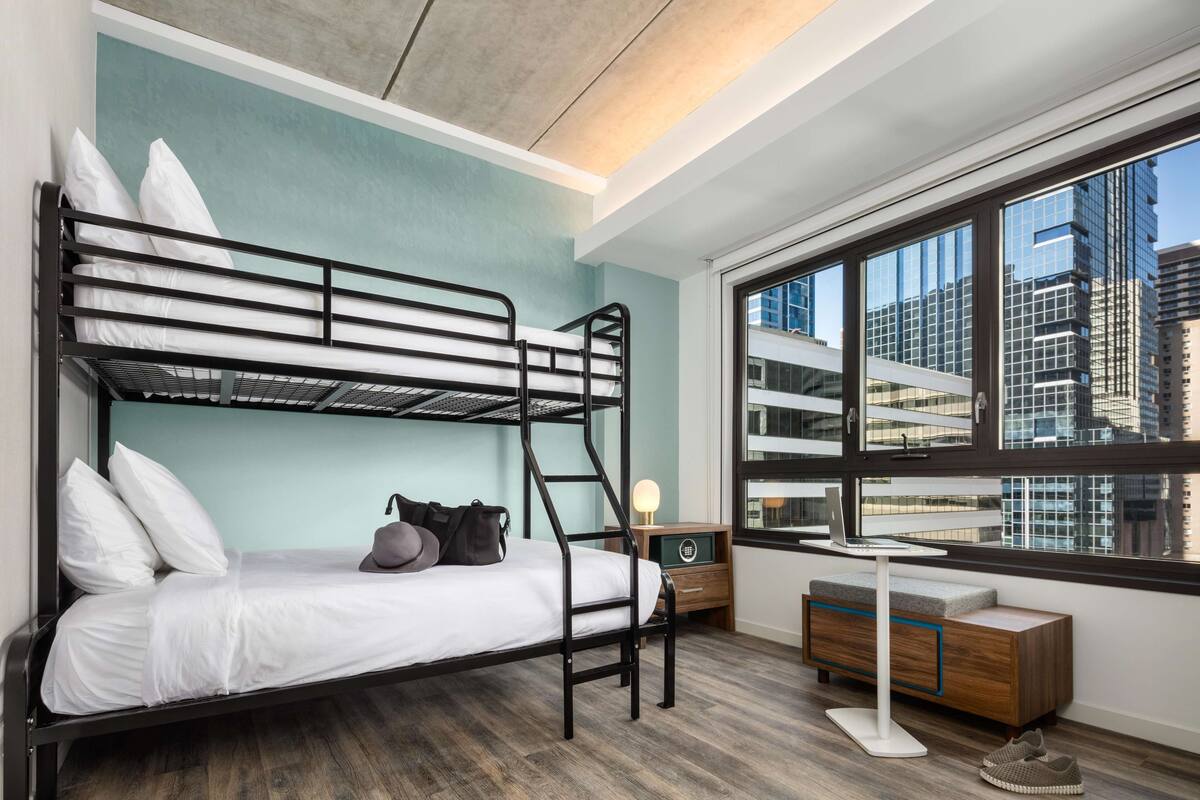 A modern hotel room with a bunk bed, a desk with a chair, a small cabinet, and a large window offering a city view.