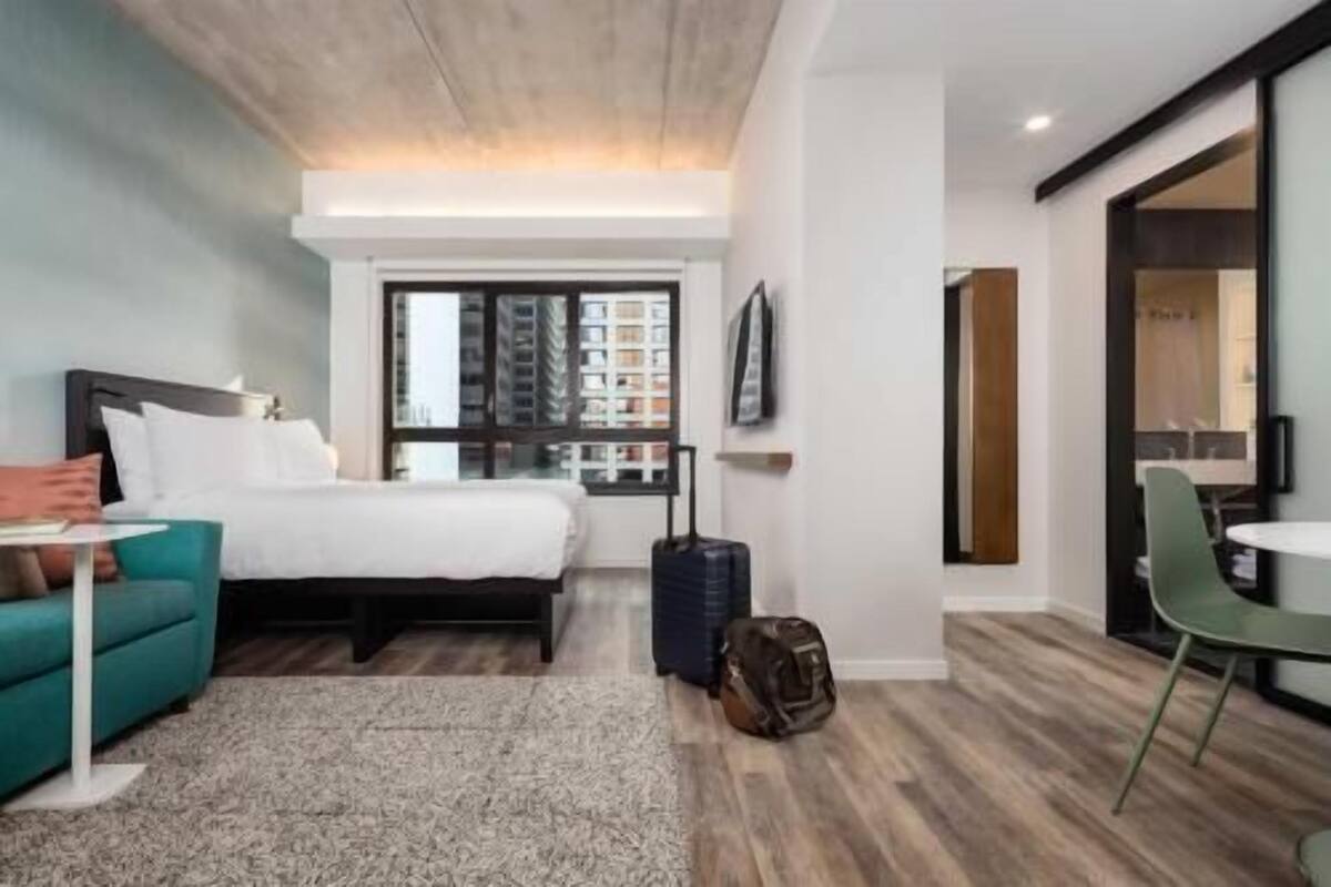 A hotel room with a bed, a desk, a chair, a suitcase, and a window with a city view.