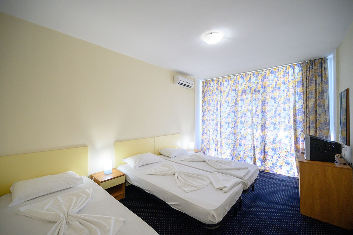 A hotel room with two beds, a nightstand, a TV, and a window with floral curtains.