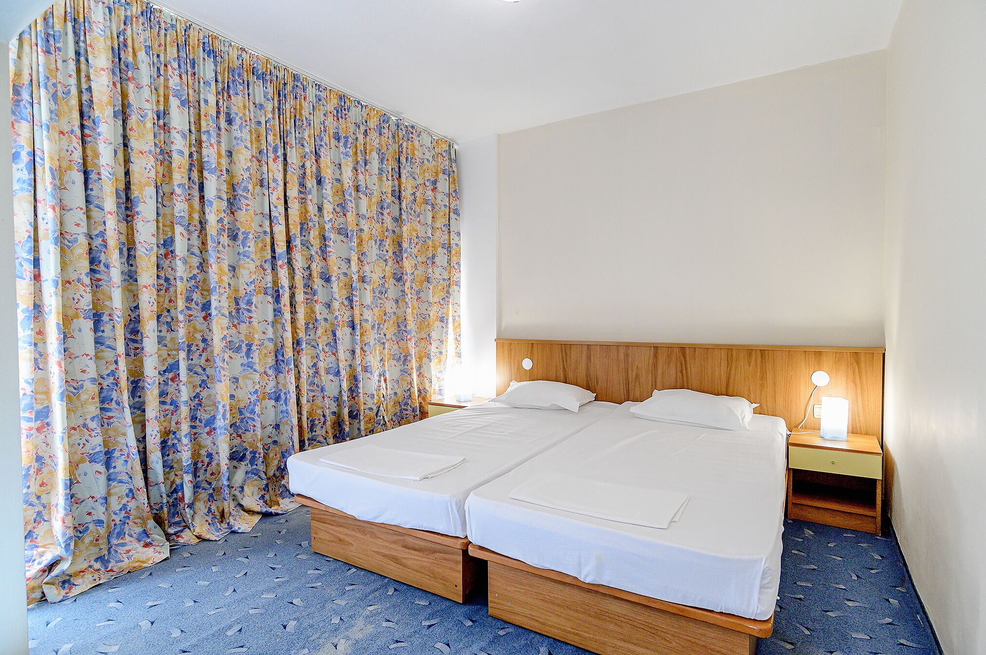 Una cama doble con cabecera de madera, una mesita de noche con lámpara y una cortina estampada con motivos florales.