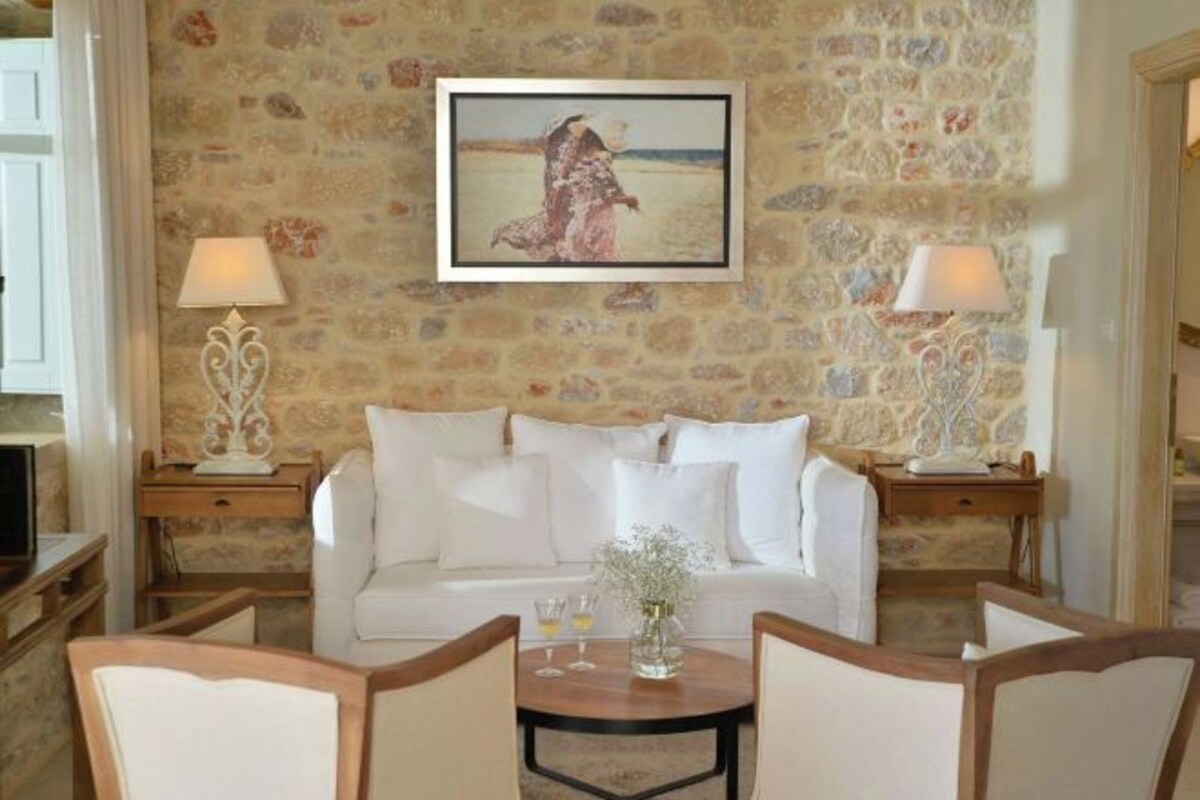 A room with a stone wall, a white sofa, two lamps, a table with a vase, and a framed picture above the sofa.