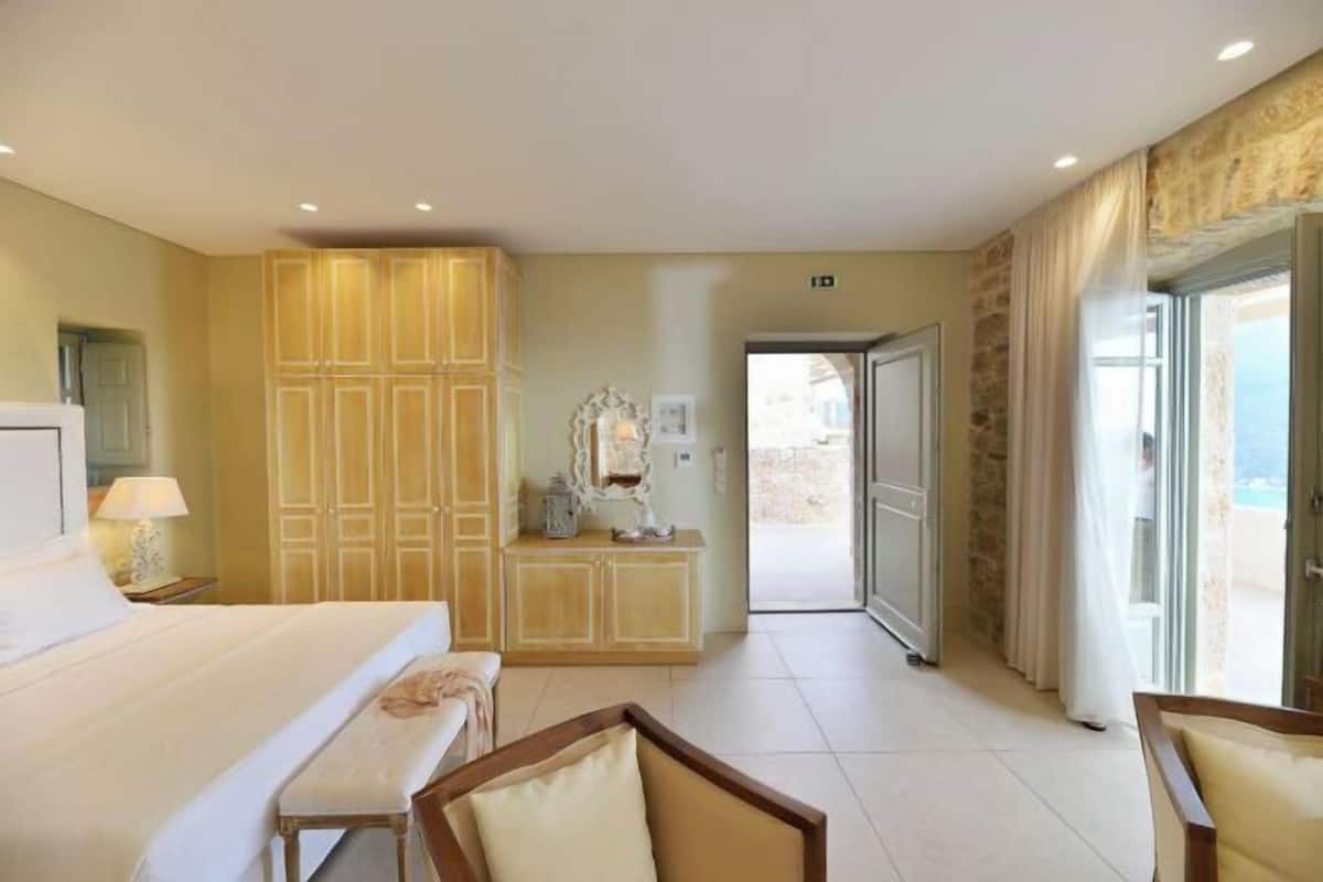 Habitación de hotel con cama, banca, tocador con espejo y vista a través de una puerta abierta.