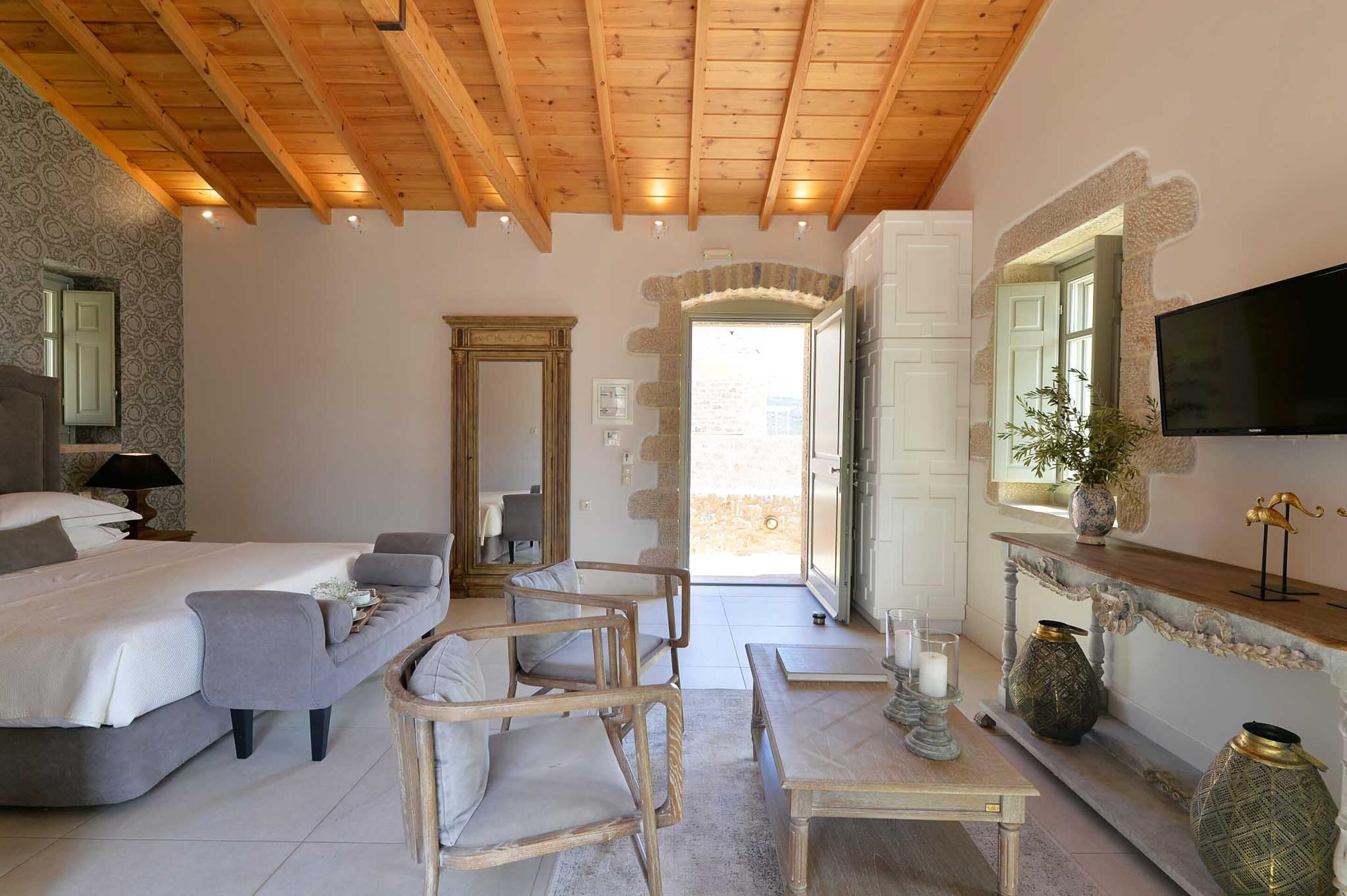 A bedroom with a bed, two chairs, a table, a TV, and a stone wall.