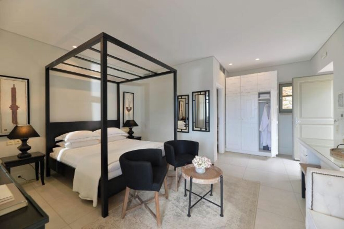 Un dormitorio moderno con cama con dosel, dos sillas negras, una mesita redonda y un baño blanco visible a través de una puerta abierta.