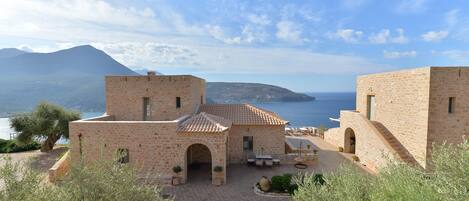 Un bâtiment en pierre doté d’une cour, d’un espace extérieur aménagé pour s’asseoir et offrant une vue sur la mer et les montagnes.