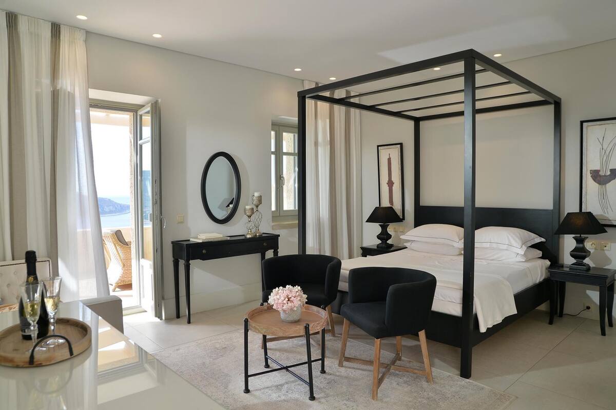 Un dormitorio moderno con una cama con dosel, una mesita de noche con lámpara, una pequeña mesa redonda con un jarrón de flores y vistas al exterior a través de un amplio ventanal.