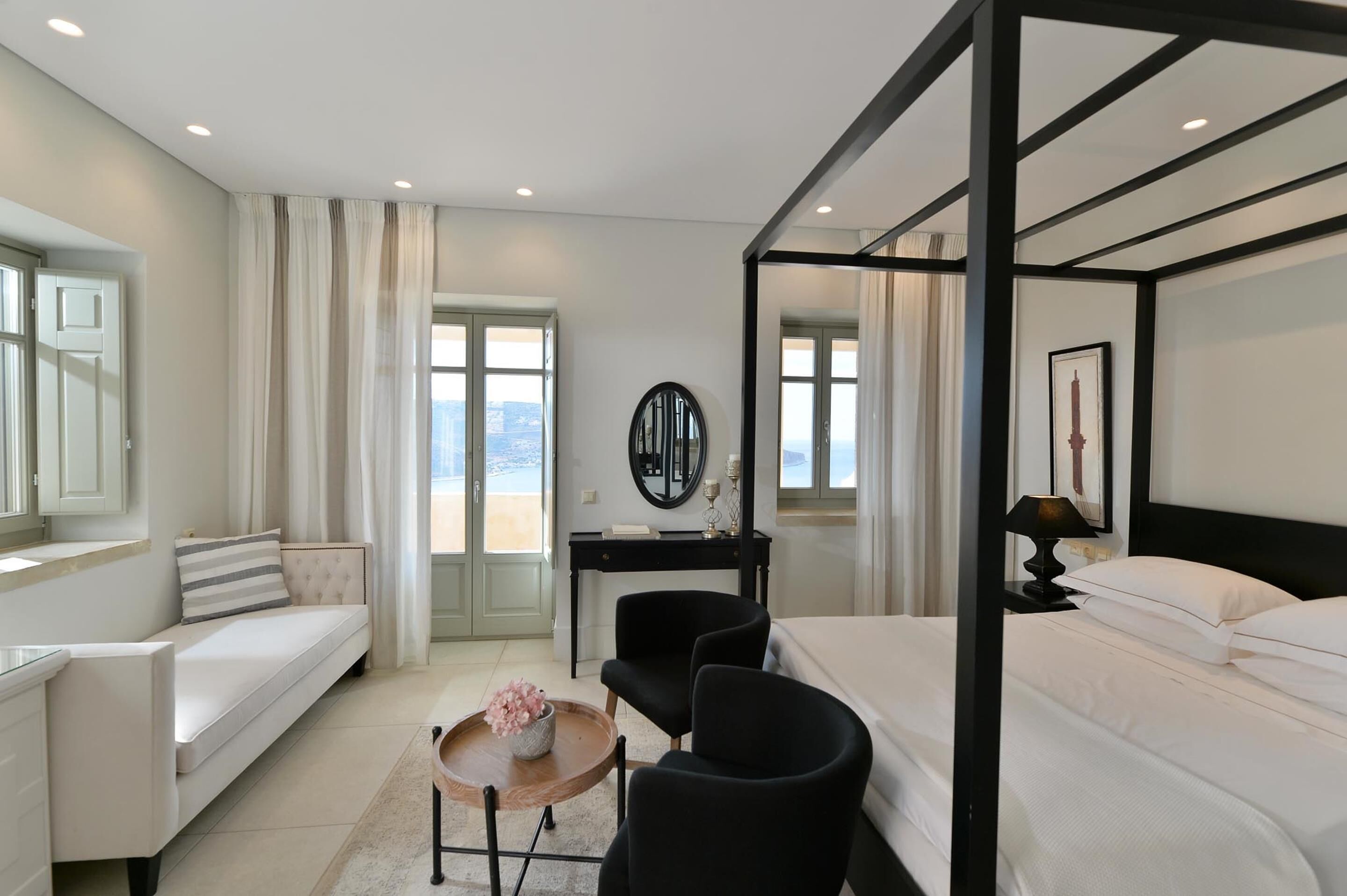 A modern hotel room with a bed, a desk, a sofa, and a view of the city.